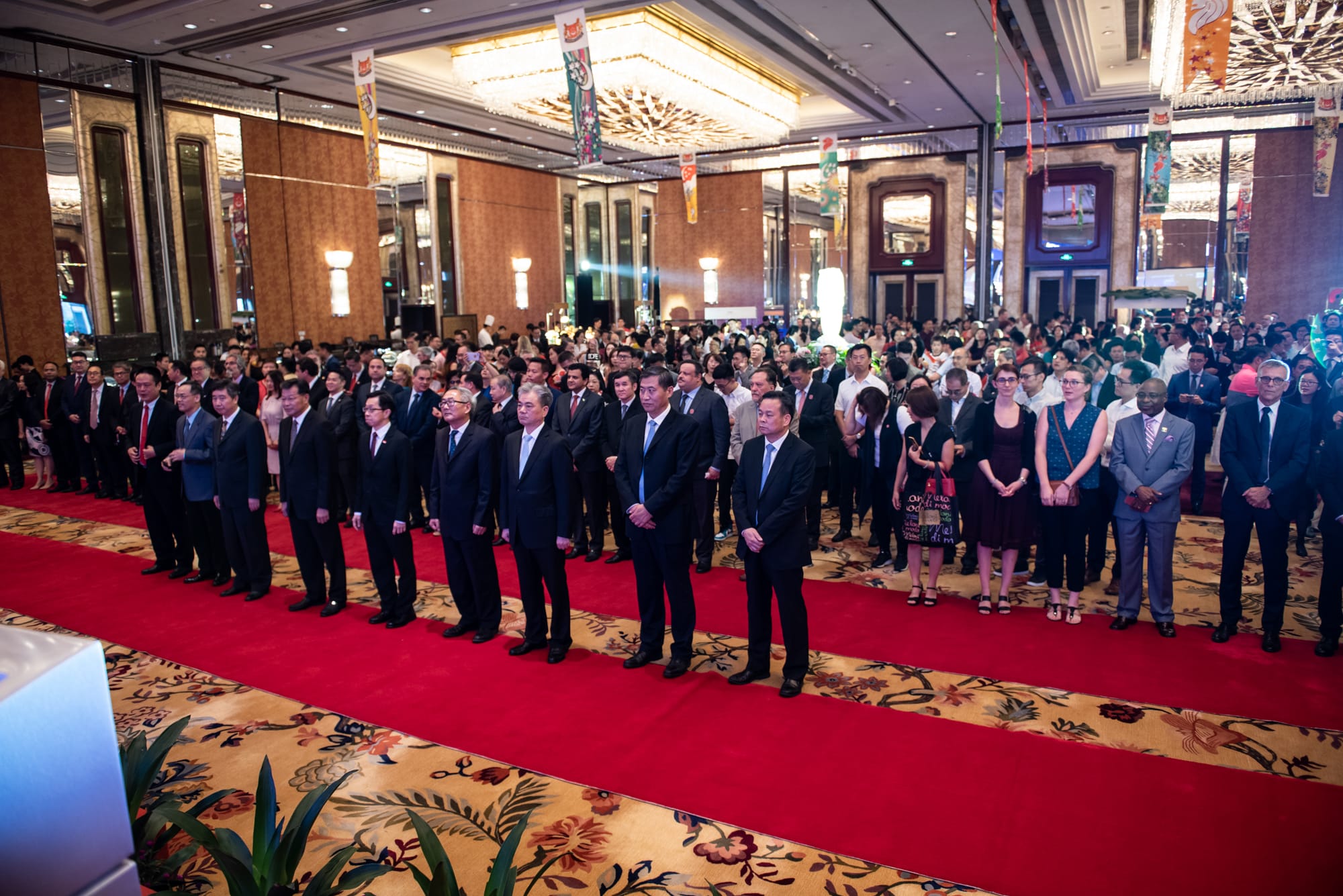A large group of people stand in a formal indoor space with a red carpet.
